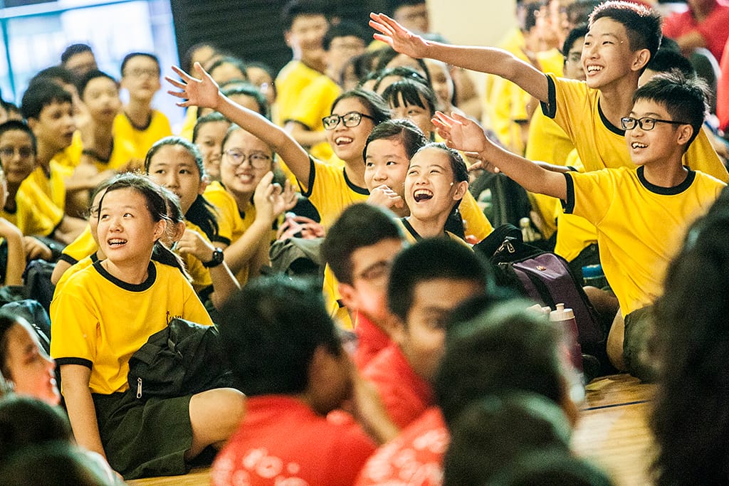 A large group of smiling children in yellow and black uniforms sit together, some raising their hands and laughing enthusiastically.